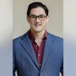 Smiling man wearing glasses and a blue blazer over a maroon shirt against a neutral background.
