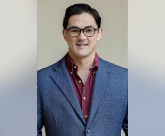 Smiling man wearing glasses and a blue blazer over a maroon shirt against a neutral background.