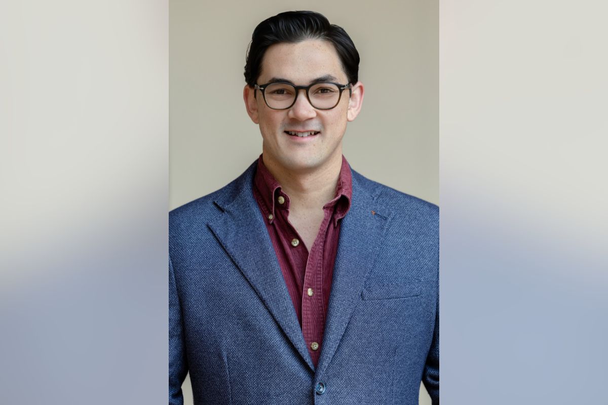 Smiling man wearing glasses and a blue blazer over a maroon shirt against a neutral background.
