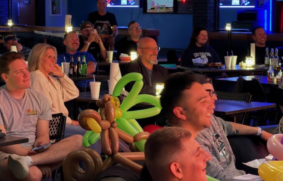 Group of people seated at tables in a bar, smiling and watching screens, with a balloon centerpiece in the foreground as decorations.”