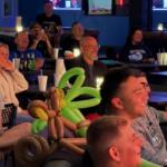 Group of people seated at tables in a bar, smiling and watching screens, with a balloon centerpiece in the foreground as decorations.”