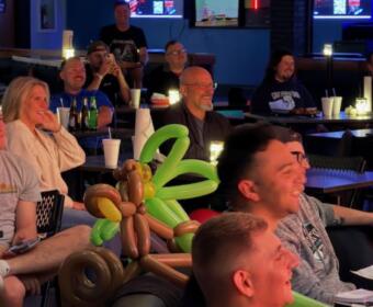 Group of people seated at tables in a bar, smiling and watching screens, with a balloon centerpiece in the foreground as decorations.”
