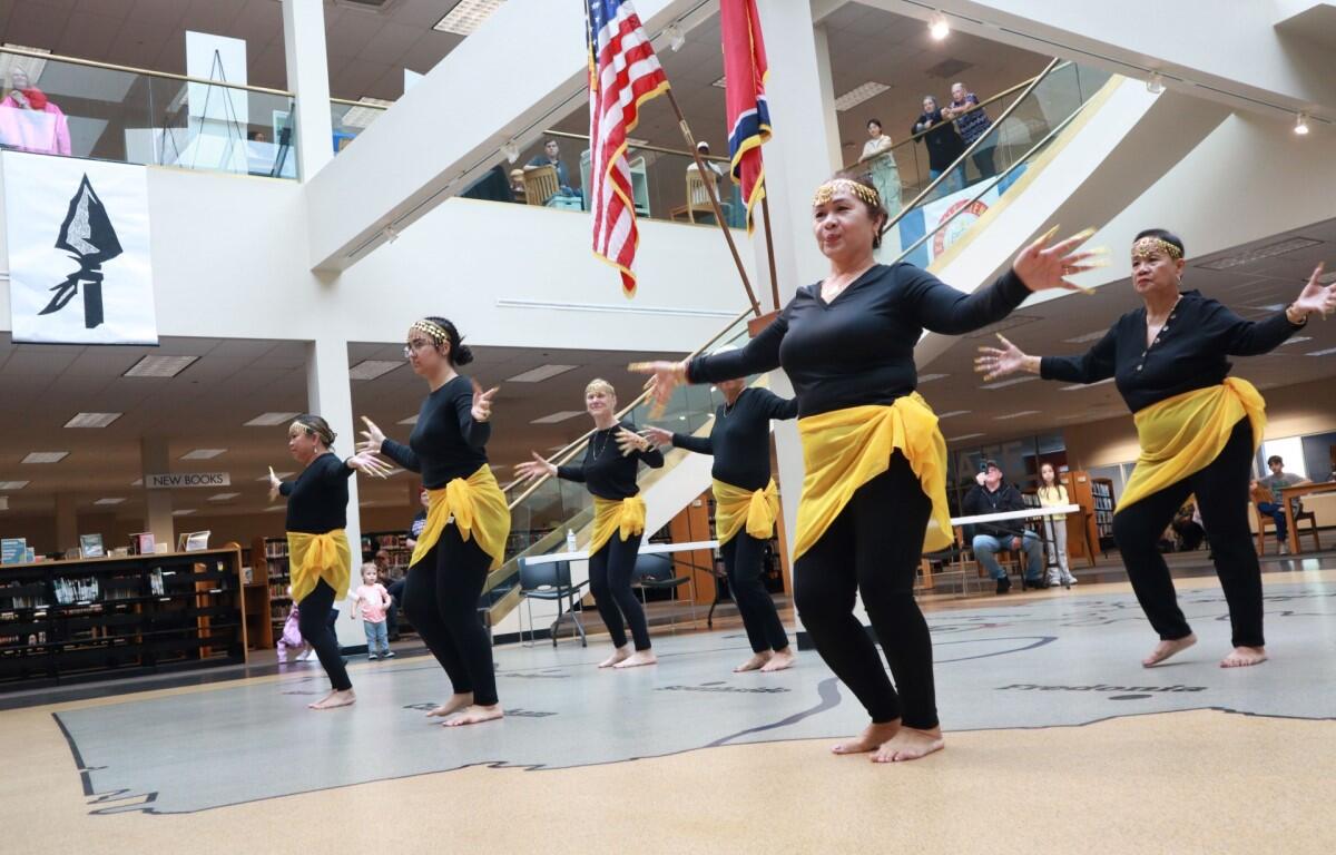 Dancers in black outfits with yellow sashes perform barefoot in a library atrium, arms outstretched as flags hang overhead.