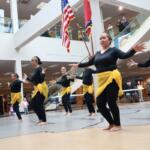 Dancers in black outfits with yellow sashes perform barefoot in a library atrium, arms outstretched as flags hang overhead.