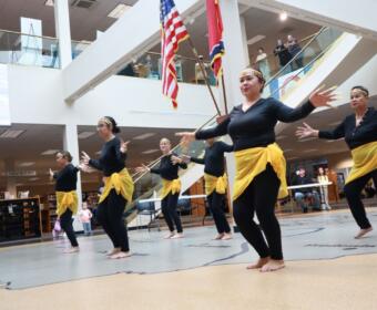 Dancers in black outfits with yellow sashes perform barefoot in a library atrium, arms outstretched as flags hang overhead.