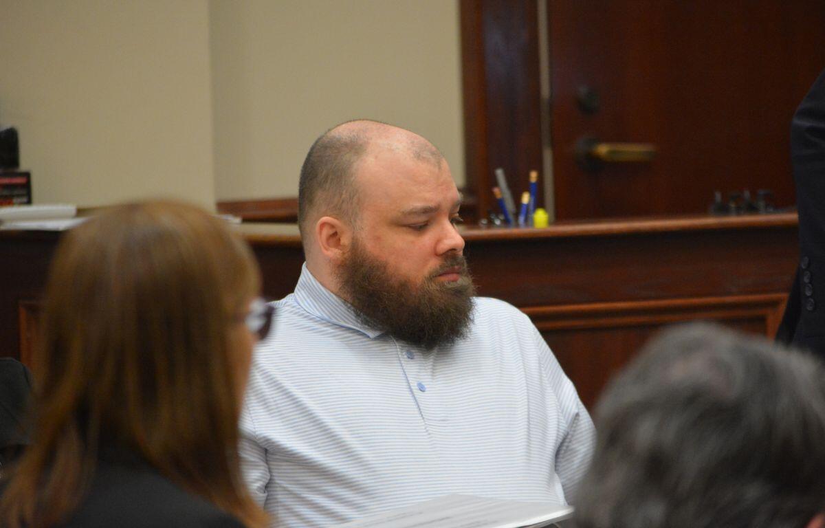 Bearded man in a light blue striped polo seated at a table in a courtroom, looking to the left.