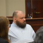 Bearded man in a light blue striped polo seated at a table in a courtroom, looking to the left.