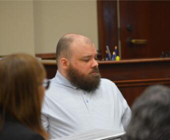 Bearded man in a light blue striped polo seated at a table in a courtroom, looking to the left.