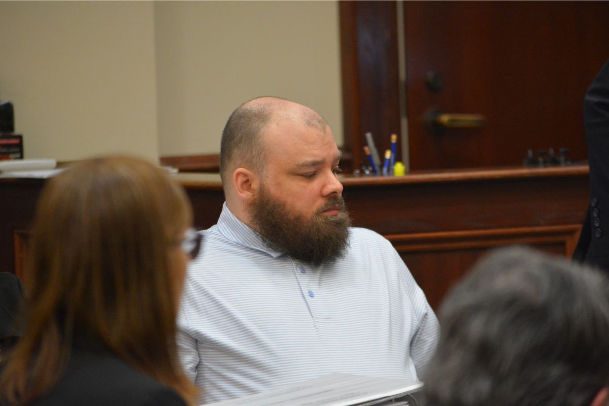 Bearded man in a light blue striped polo seated at a table in a courtroom, looking to the left.
