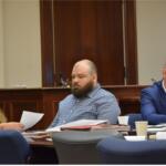 Three adults sit at a wooden conference table reviewing papers in a formal meeting room. The woman on the left wears glasses and looks at documents, the bearded man in the middle glances downward, and a bald man in a blue suit sits on the right with papers in front of him.