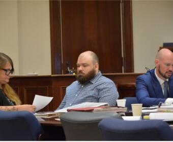 Three adults sit at a wooden conference table reviewing papers in a formal meeting room. The woman on the left wears glasses and looks at documents, the bearded man in the middle glances downward, and a bald man in a blue suit sits on the right with papers in front of him.