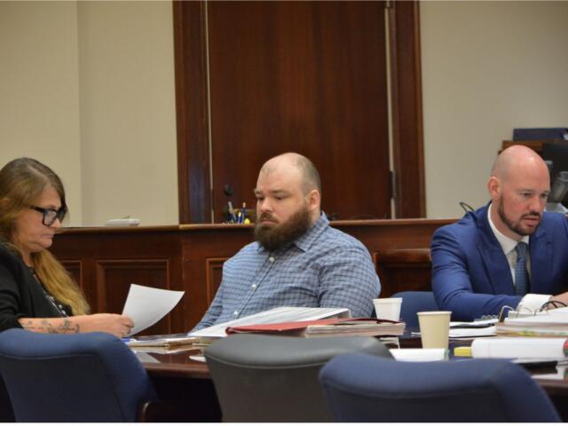 Three adults sit at a wooden conference table reviewing papers in a formal meeting room. The woman on the left wears glasses and looks at documents, the bearded man in the middle glances downward, and a bald man in a blue suit sits on the right with papers in front of him.