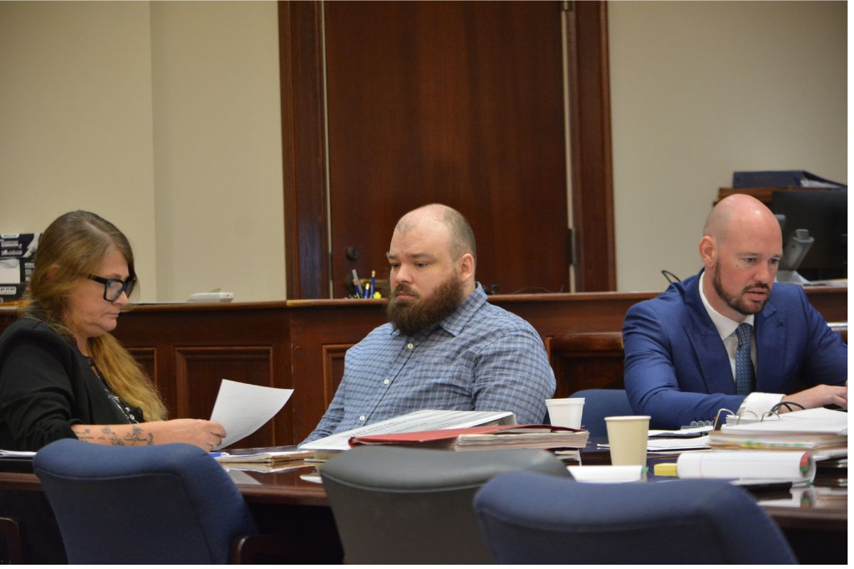 Three adults sit at a wooden conference table reviewing papers in a formal meeting room. The woman on the left wears glasses and looks at documents, the bearded man in the middle glances downward, and a bald man in a blue suit sits on the right with papers in front of him.