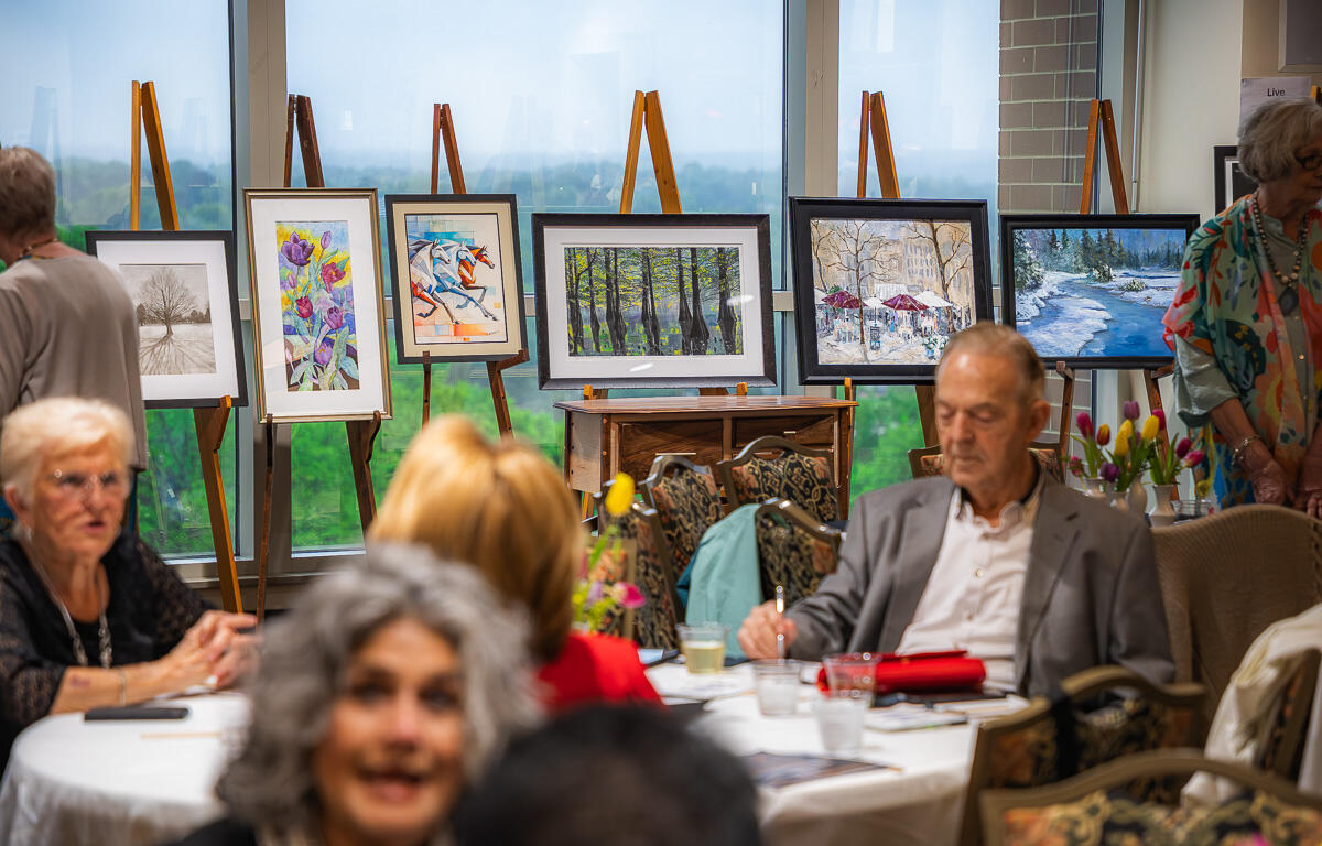 Senior adults seated around a round table at an art show, with framed paintings on easels along a large window in the background.