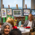 Senior adults seated around a round table at an art show, with framed paintings on easels along a large window in the background.