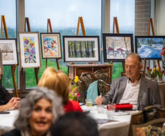 Senior adults seated around a round table at an art show, with framed paintings on easels along a large window in the background.