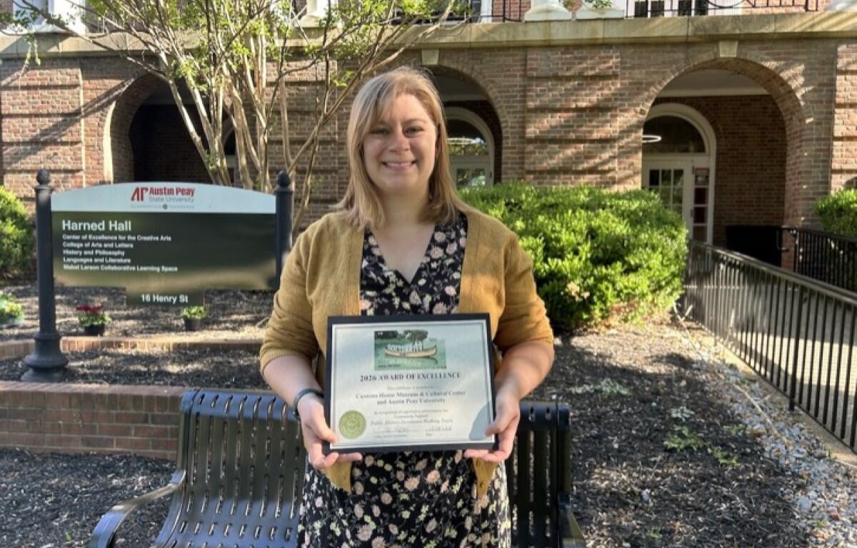 Smiling woman in a mustard cardigan holding an award plaque outdoors in front of a brick building and a bench.