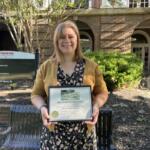 Smiling woman in a mustard cardigan holding an award plaque outdoors in front of a brick building and a bench.