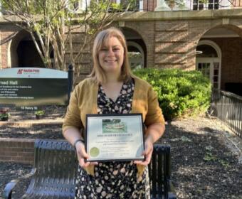 Smiling woman in a mustard cardigan holding an award plaque outdoors in front of a brick building and a bench.