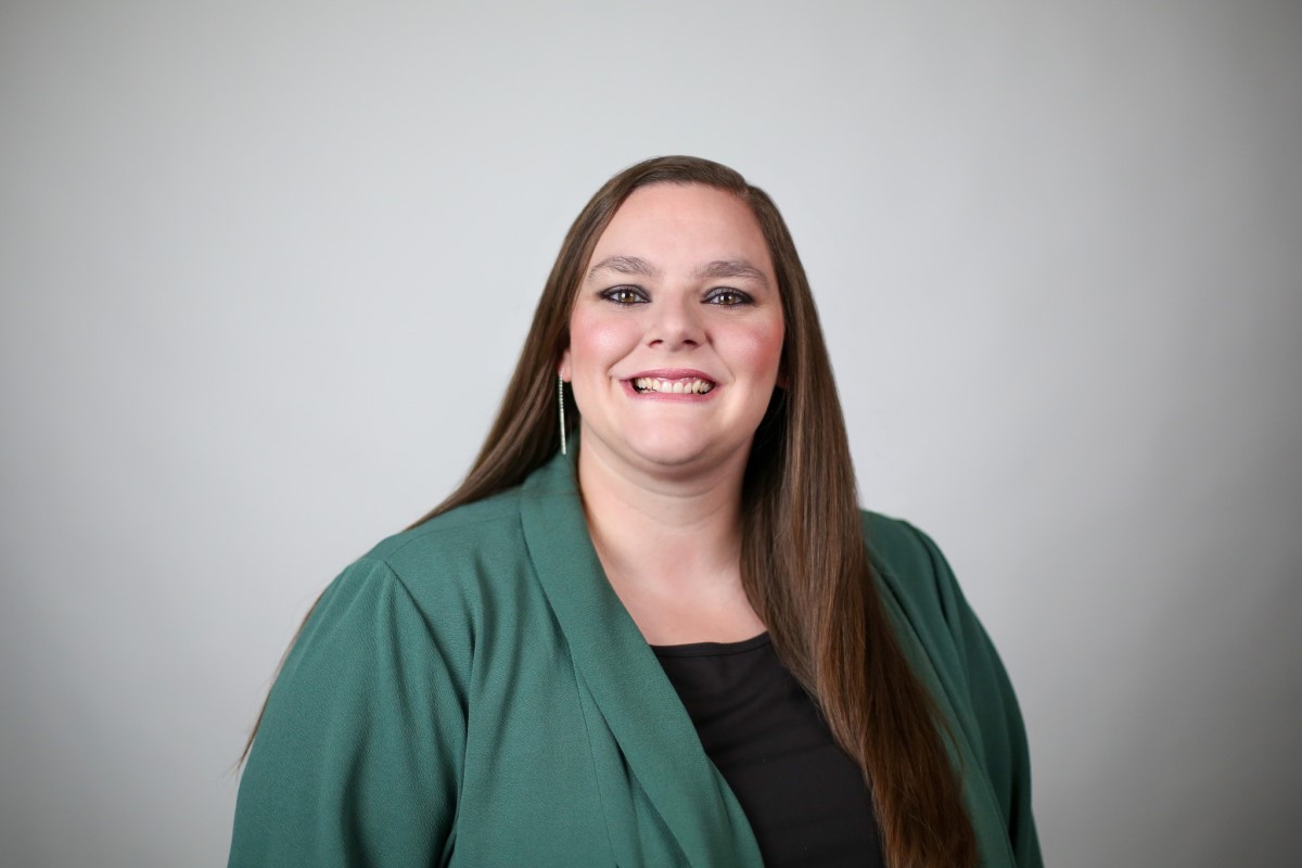 Professional headshot of a smiling woman with long brown hair, wearing a green blazer and black top, against a neutral backdrop