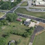 Aerial view of a busy multi-lane intersection with green fields and a few houses nearby, plus a large building with a parking lot to the top right.
