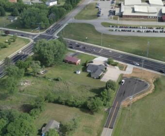 Aerial view of a busy multi-lane intersection with green fields and a few houses nearby, plus a large building with a parking lot to the top right.
