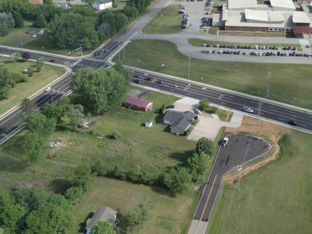Aerial view of a busy multi-lane intersection with green fields and a few houses nearby, plus a large building with a parking lot to the top right.