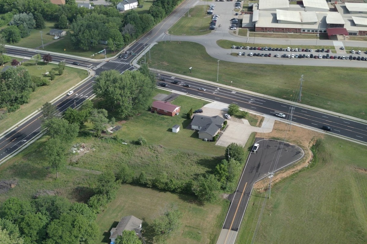 Aerial view of a busy multi-lane intersection with green fields and a few houses nearby, plus a large building with a parking lot to the top right.
