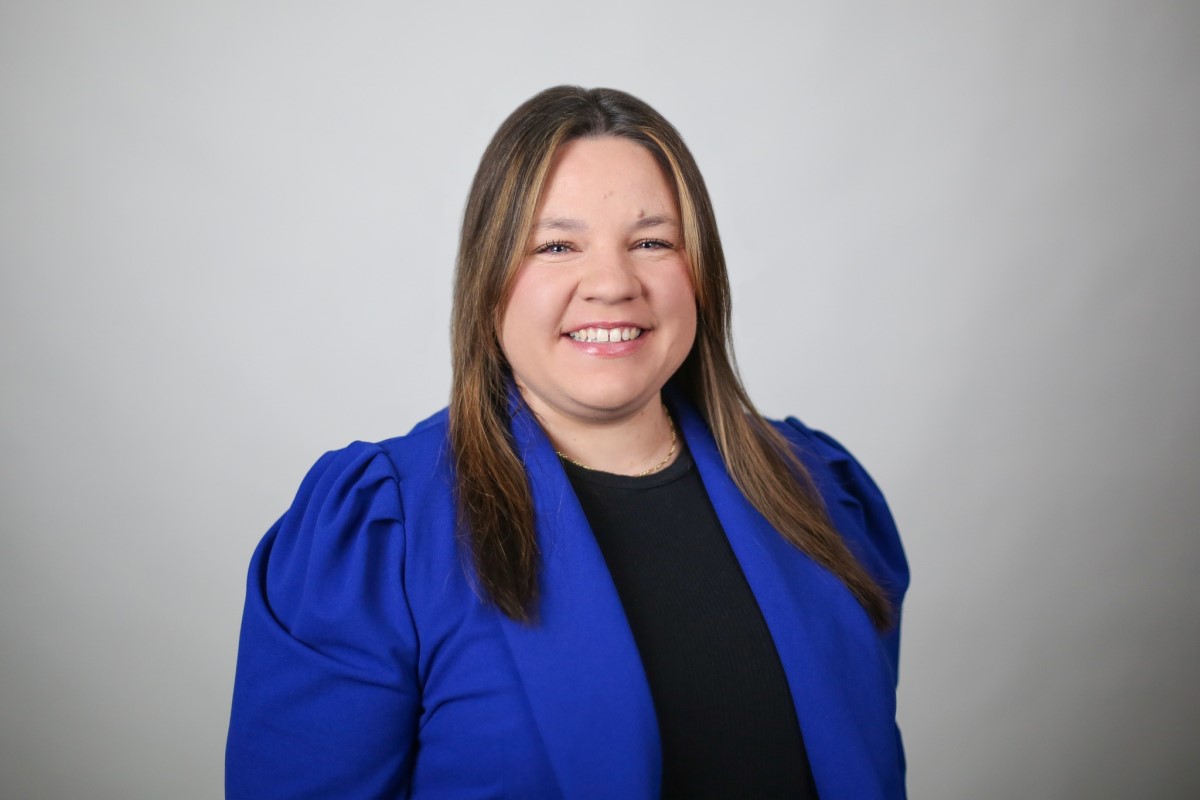 Smiling woman with shoulder-length brown hair wearing a royal blue blazer over a black top, against a light gray background.