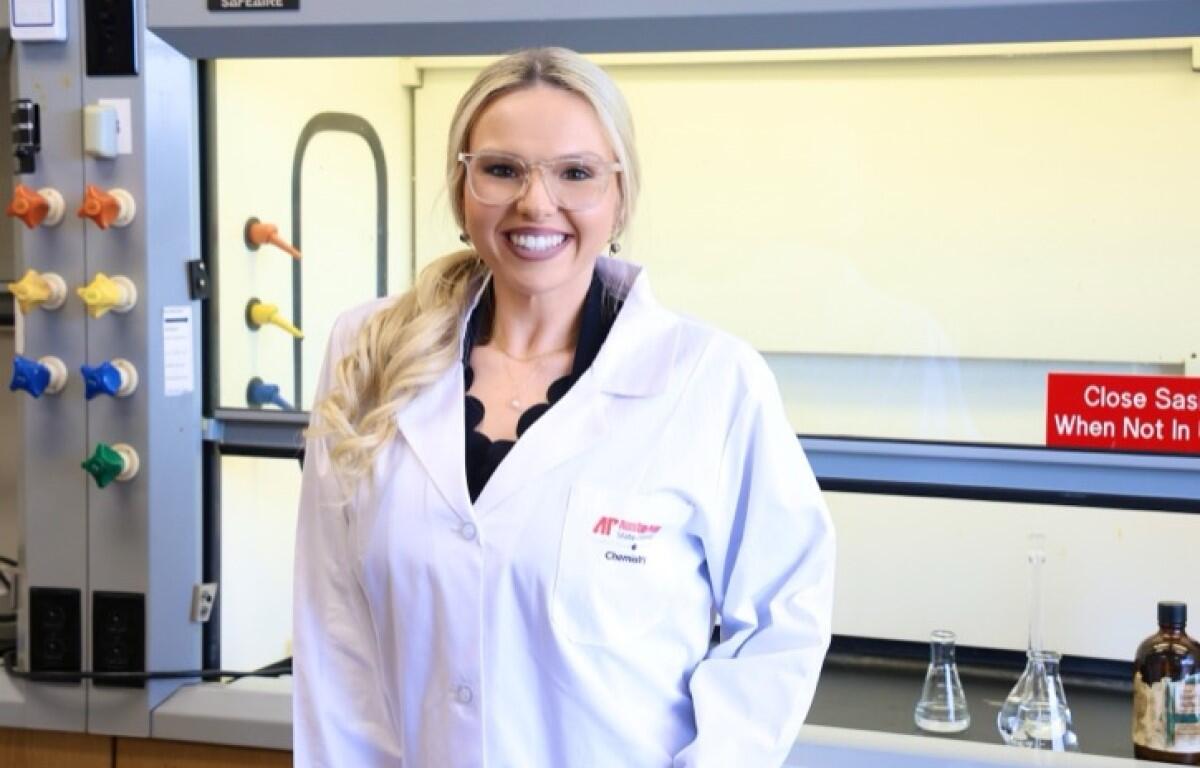 Woman in a white lab coat and safety glasses smiling in a laboratory with glassware and equipment in the background.