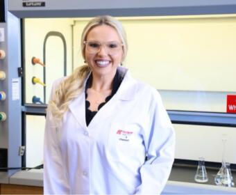 Woman in a white lab coat and safety glasses smiling in a laboratory with glassware and equipment in the background.