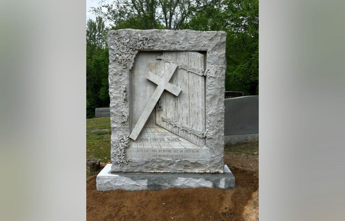 Gravestone with a diagonal cross leaning against a carved door, framed by floral reliefs and an inscription at the base.