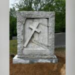 Gravestone with a diagonal cross leaning against a carved door, framed by floral reliefs and an inscription at the base.