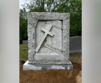 Gravestone with a diagonal cross leaning against a carved door, framed by floral reliefs and an inscription at the base.