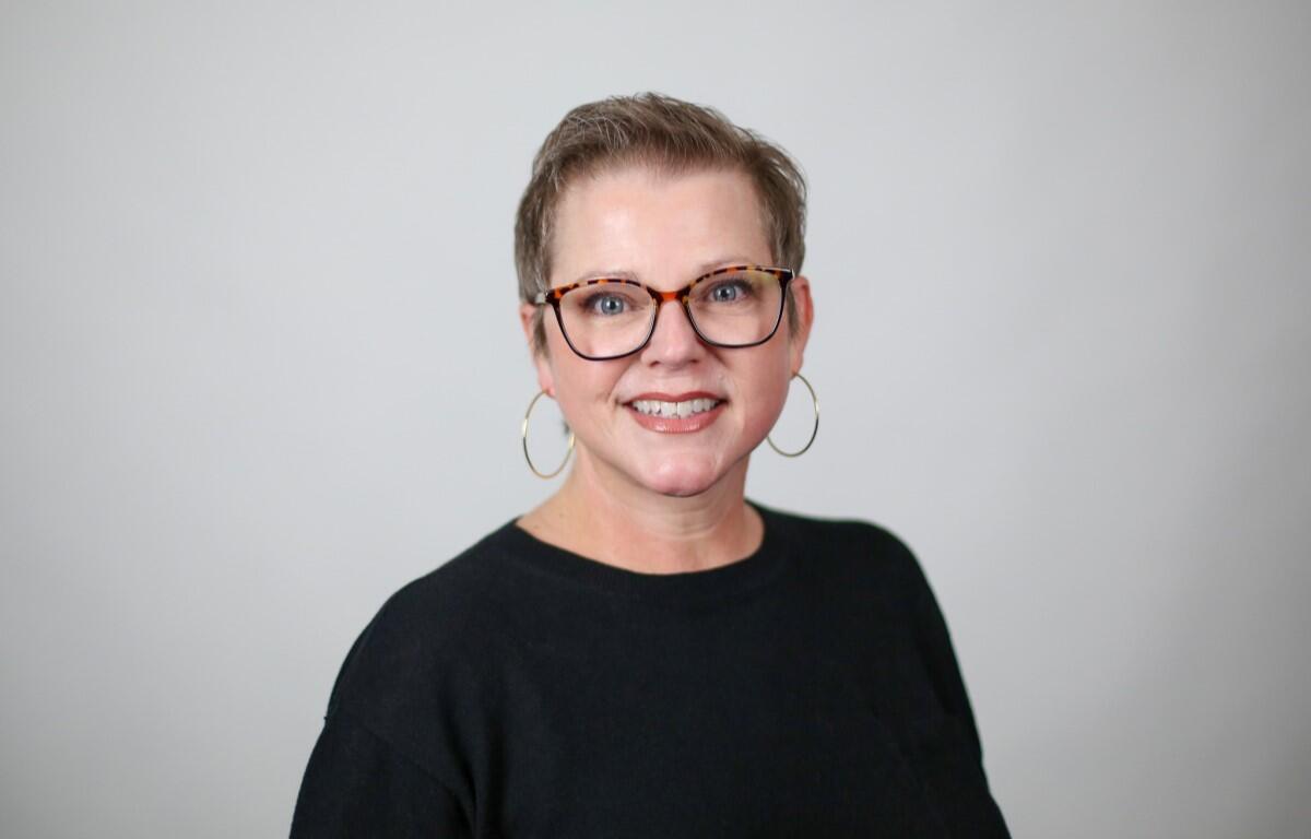 Smiling woman with short hair and tortoiseshell glasses, wearing a black top and gold hoop earrings, against a light gray background.