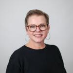 Smiling woman with short hair and tortoiseshell glasses, wearing a black top and gold hoop earrings, against a light gray background.