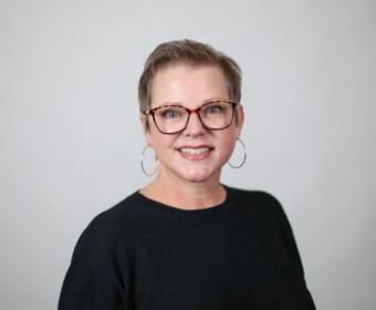 Smiling woman with short hair and tortoiseshell glasses, wearing a black top and gold hoop earrings, against a light gray background.