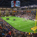 Indoor football arena with a full crowd, players on a green turf field, and a large hanging scoreboard showing game stats and time.