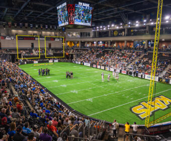 Indoor football arena with a full crowd, players on a green turf field, and a large hanging scoreboard showing game stats and time.