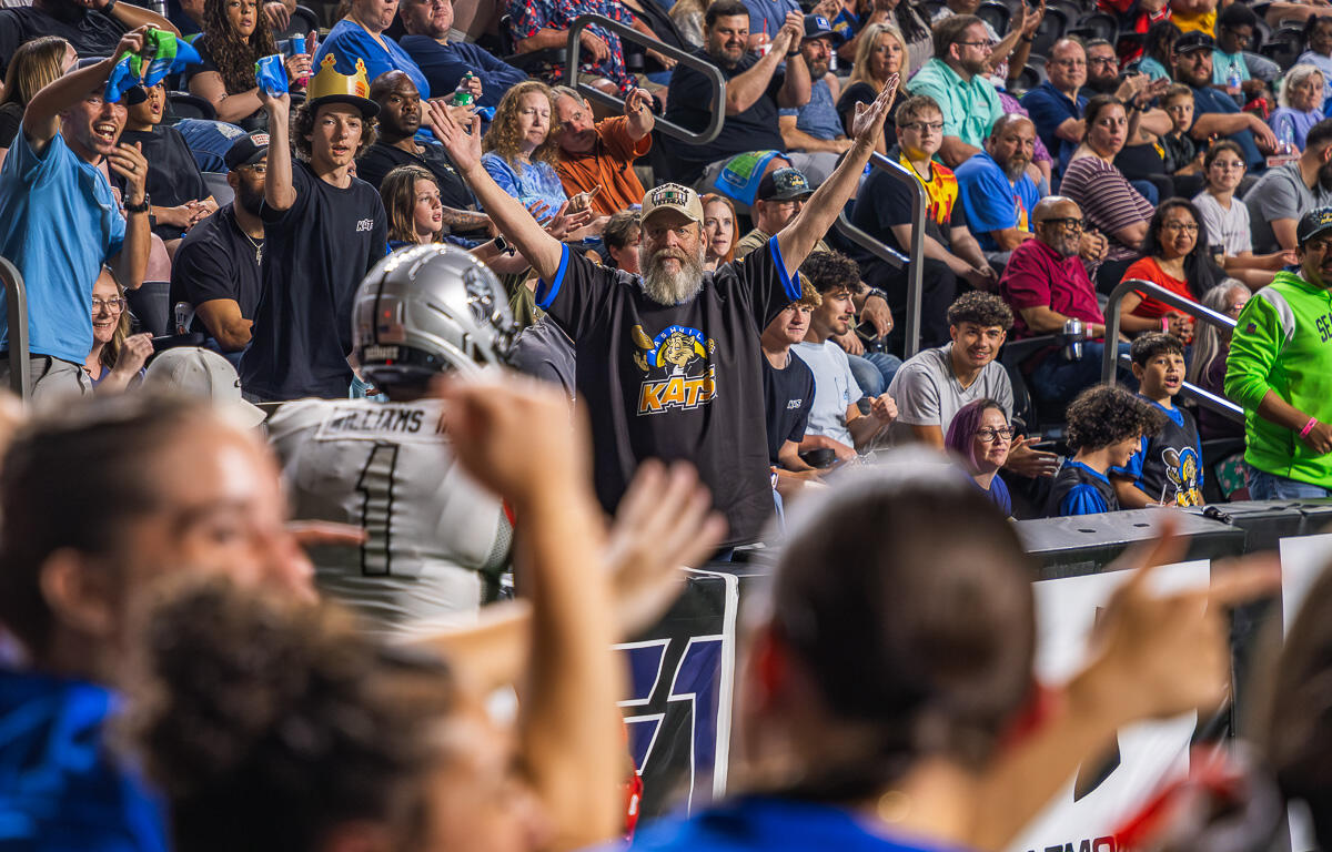 Bearded man in a black KATs shirt raises both arms amid cheering crowd at a football stadium.