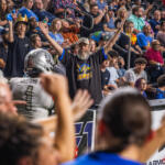 Bearded man in a black KATs shirt raises both arms amid cheering crowd at a football stadium.