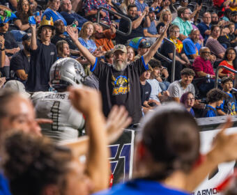 Bearded man in a black KATs shirt raises both arms amid cheering crowd at a football stadium.