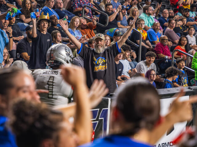 Bearded man in a black KATs shirt raises both arms amid cheering crowd at a football stadium.