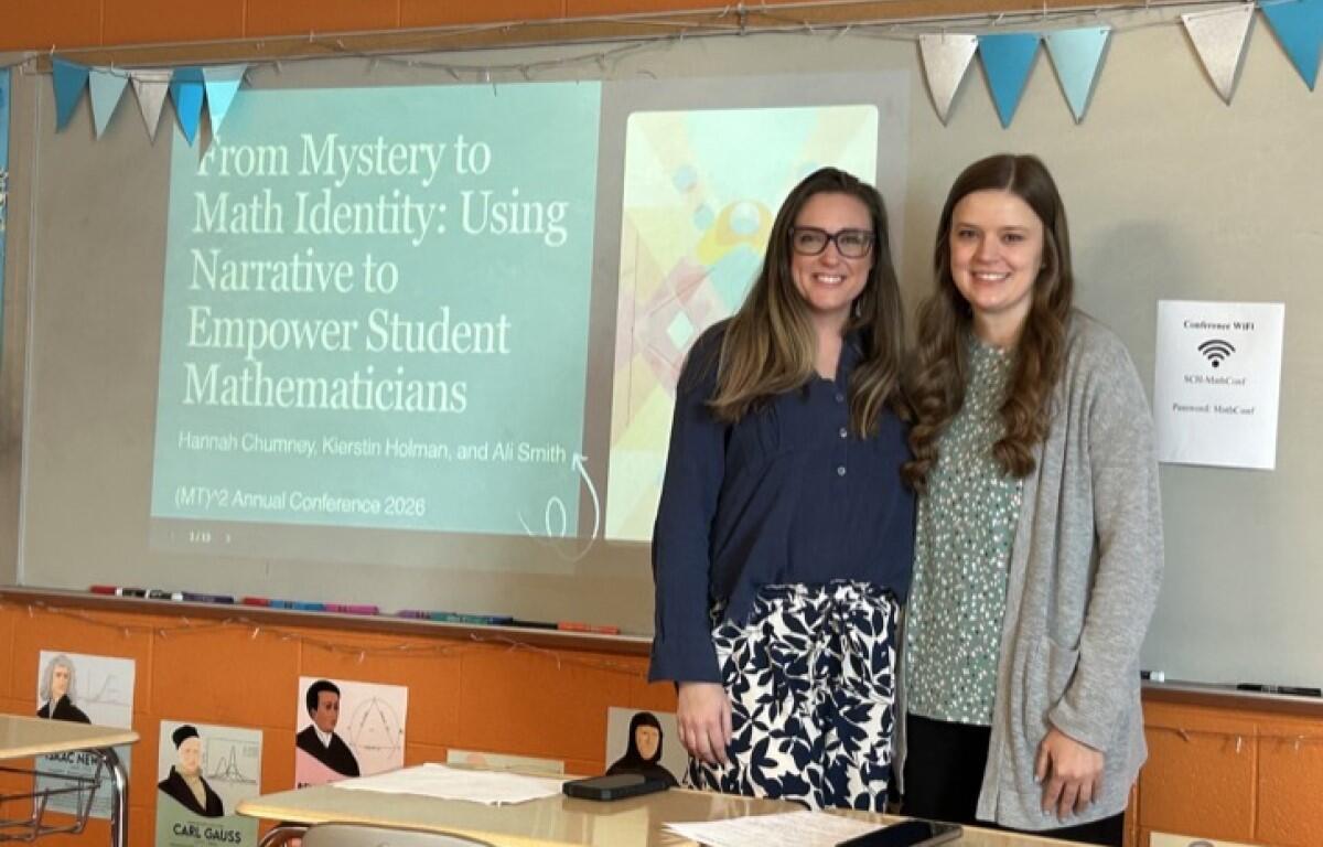 Two women pose in a classroom beside a projected slide about empowering student mathematicians in math identity research.