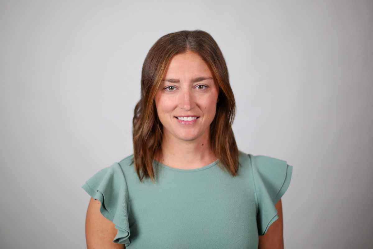 Head-and-shoulders portrait of a smiling woman with brown hair in a green short-sleeve top, neutral backdrop.