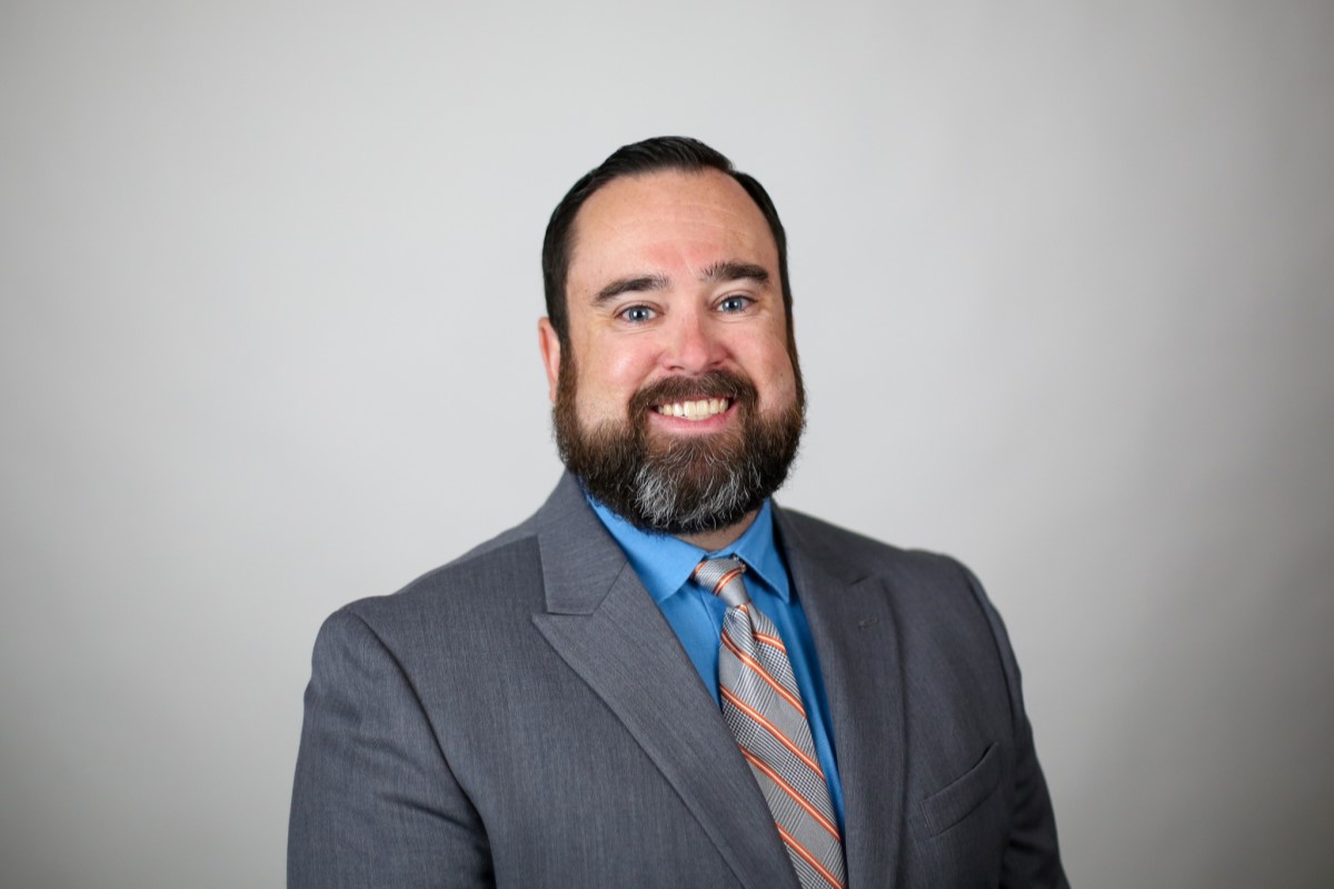 Smiling man in a gray suit and blue shirt with a striped tie against a neutral backdrop, posing for a professional headshot.