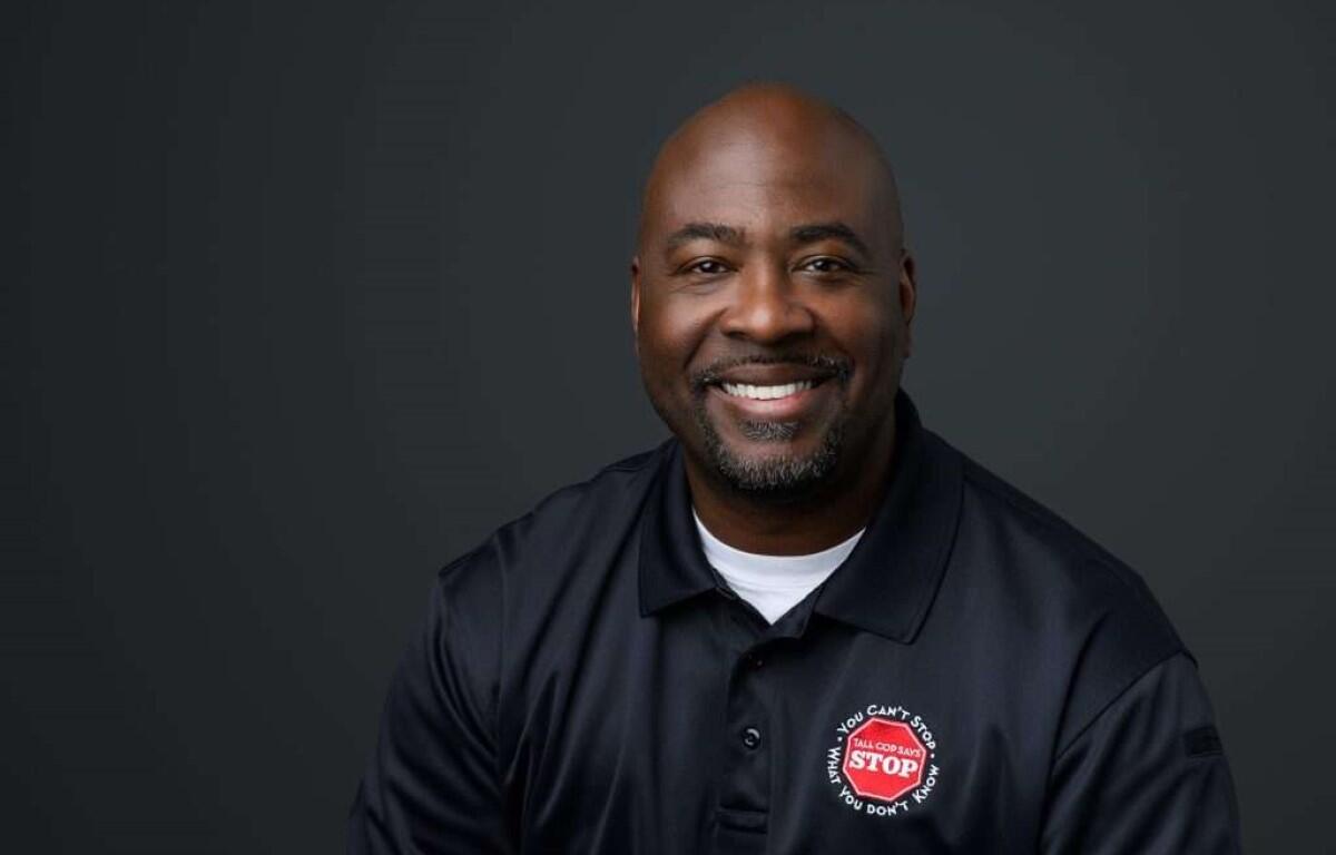 Smiling African American man in a navy polo shirt with a red STOP patch, against a dark studio background (head-and-shoulders portrait).
