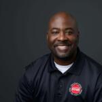 Smiling African American man in a navy polo shirt with a red STOP patch, against a dark studio background (head-and-shoulders portrait).