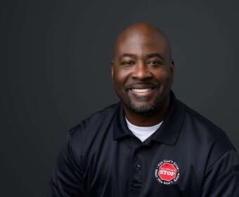 Smiling African American man in a navy polo shirt with a red STOP patch, against a dark studio background (head-and-shoulders portrait).