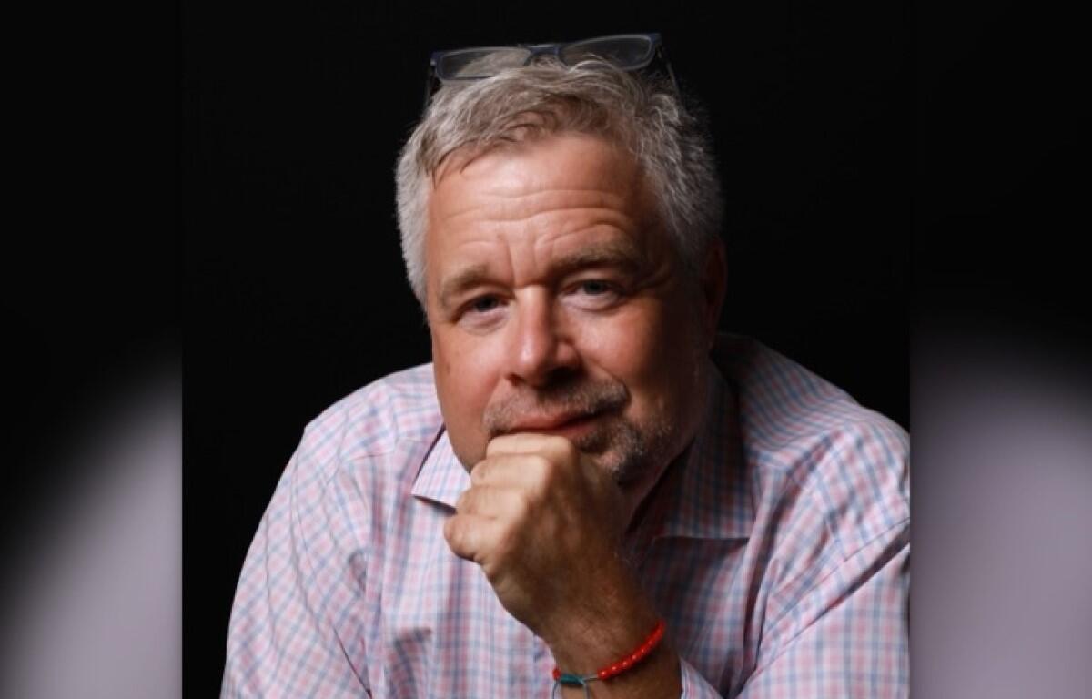 Portrait of a middle-aged man with gray hair in a pink checkered shirt, resting his chin on his hand.